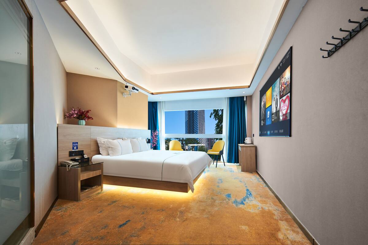 Quarto de hotel moderno com uma cama grande, uma escrivaninha com luminária, vista para a cidade e televisão na parede.