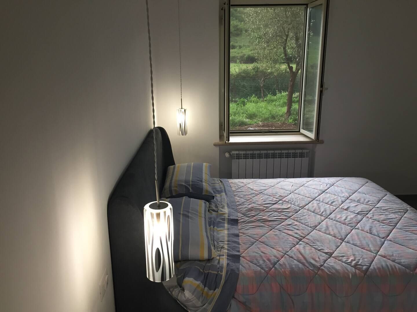 A bedroom with a bed, a hanging light fixture, a window with a view of greenery, and a radiator.