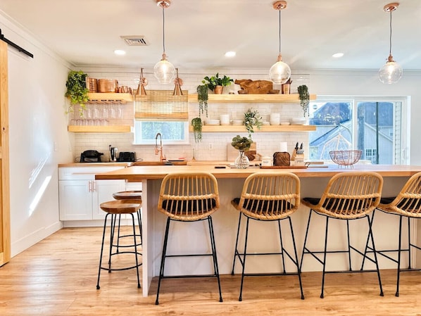 Een keuken met een centraal eiland, barstoelen, hanglampen en planten op planken.