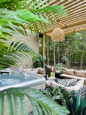 Una vasca idromassaggio sotto una pergola in legno con una lampada pendente, circondata da divani e vegetazione lussureggiante.