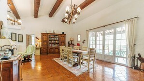 A dining area with a wooden table and chairs, a chandelier, and a view through large windows. - Villa Hokiss - Private pool on 1 ha of nature (Le Muy)