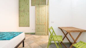 A small, well-lit room with a bed, a green chair, a wooden table, and a door.