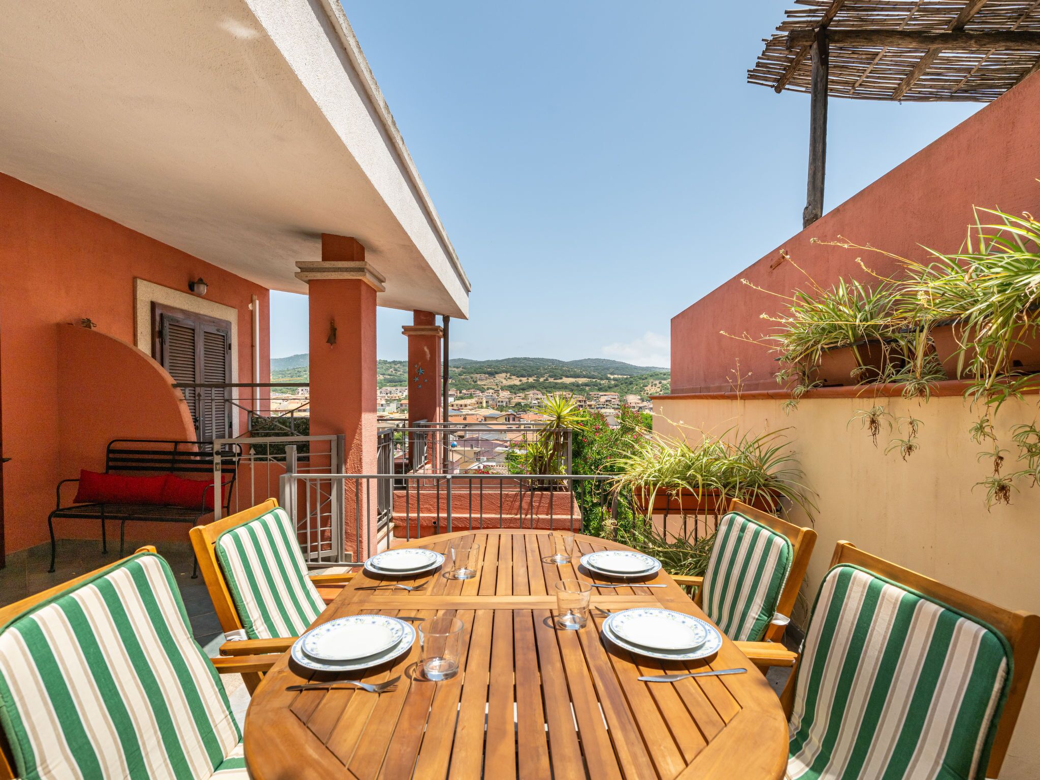 Mesa de madera preparada para cuatro personas con sillas a rayas, un banco rojo y plantas en macetas en un balcón con vista a la ciudad.