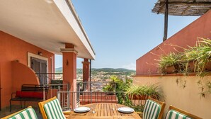 A wooden table set for four with striped chairs, a red bench, and potted plants on a balcony with a city view.