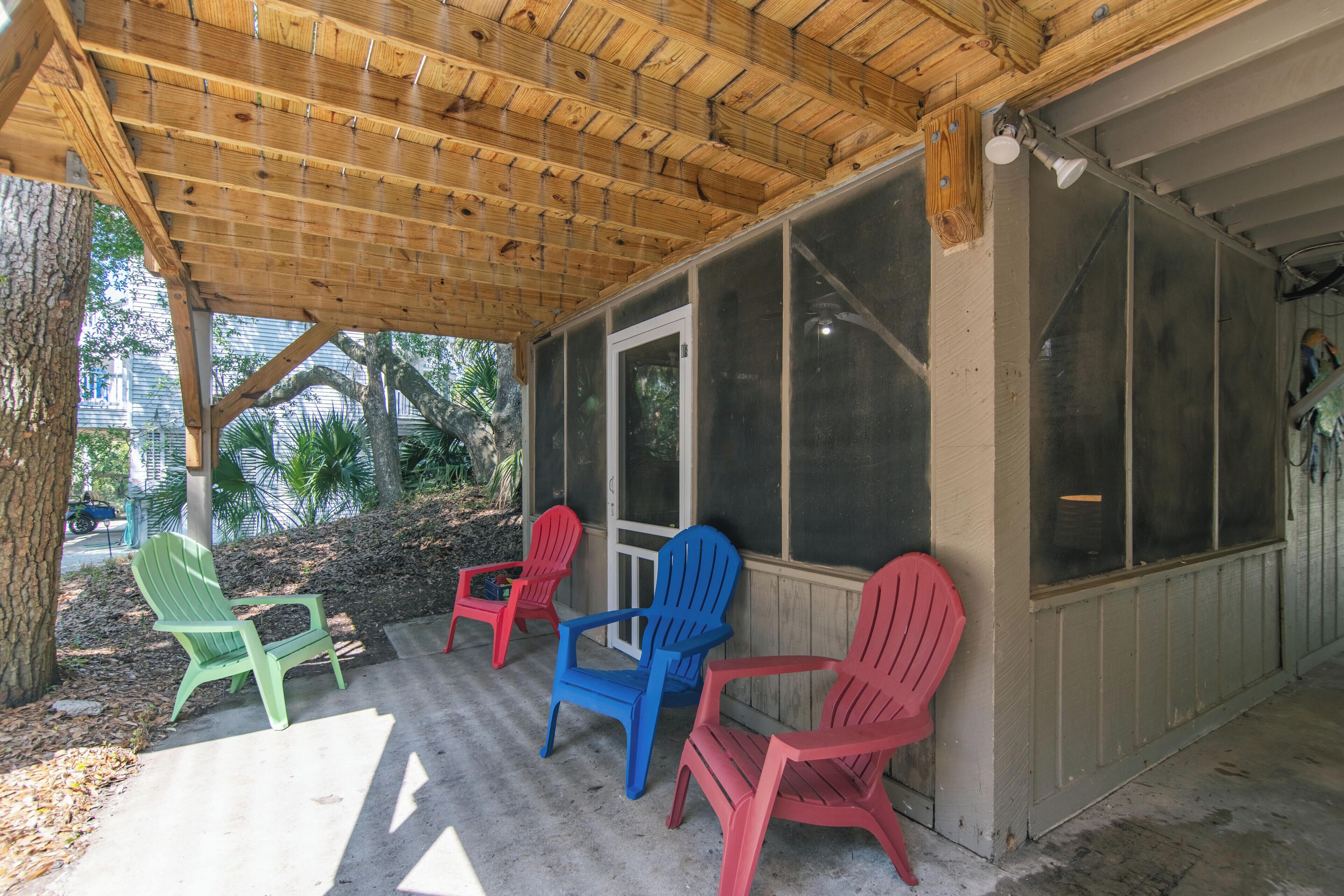 Ein überdachter Balkon mit vier Adirondack-Stühlen in Grün, Blau und Rot, einer Holzdecke und Blick auf ein Haus durch große Fenster.