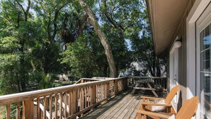 Una terraza de madera con barandilla, sillas de madera y vista a árboles y un edificio al fondo.