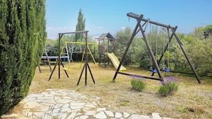 Un terrain de jeux avec des balançoires et un toboggan, entouré de verdure et bordé d’un chemin en pierre.