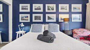 A bedroom with a large bed, multiple framed pictures on the wall, a bedside lamp, and a patterned throw blanket.