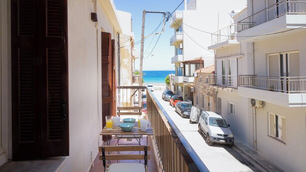 A narrow street with parked cars, a table with breakfast items, and a view of the sea.