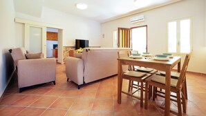A living room with a sofa, a dining table with chairs, a TV, and a kitchen area in the background.