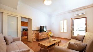 A living room with a sofa, a coffee table with a vase, a TV on a wooden cabinet, and a kitchen in the background.