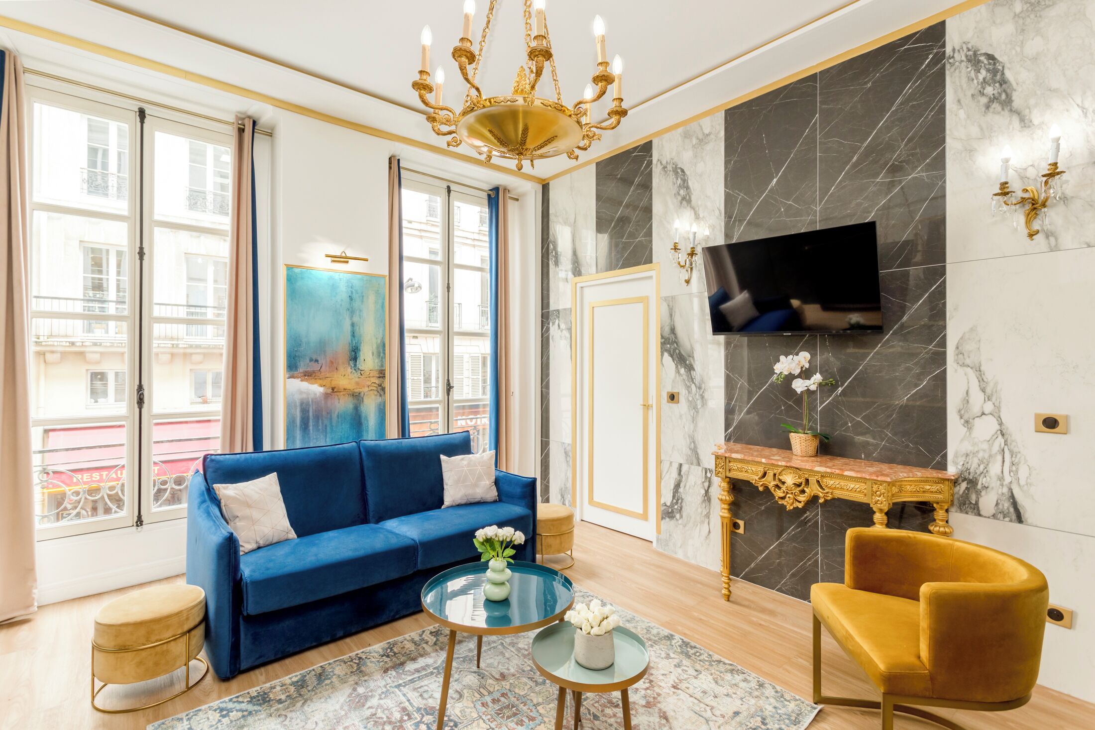 Een woonkamer met een blauwe bank, een gouden fauteuil, een ronde salontafel en een televisie bevestigd aan een marmeren muur.