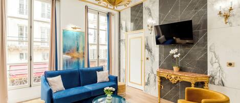Een woonkamer met een blauwe bank, een gouden fauteuil, een ronde salontafel en een televisie bevestigd aan een marmeren muur.