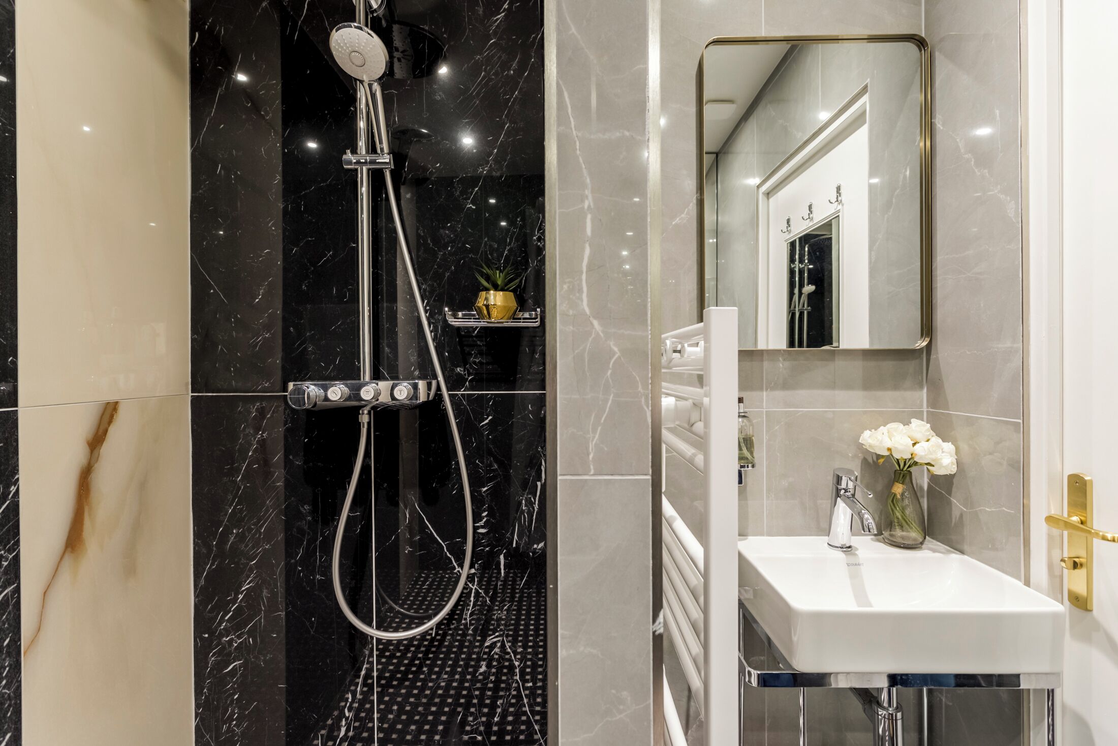 Een moderne badkamer met een zwart-wit marmeren ontwerp, een douche met handdouche, een witte wasbak met een bloemenvaas en een spiegel met gouden lijst.