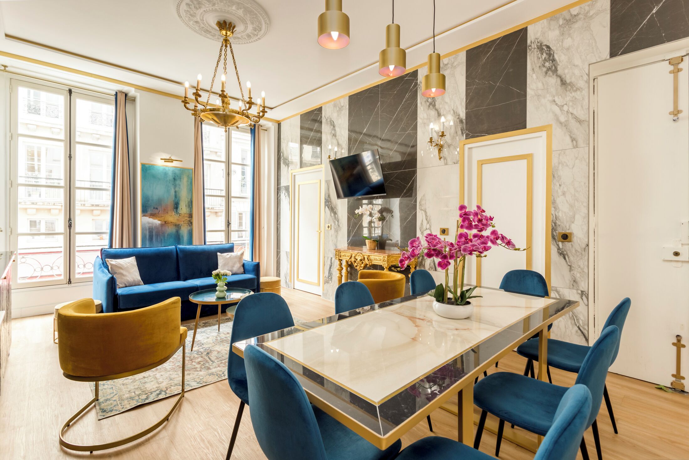 A modern living room with a blue sofa, a dining table with chairs, a chandelier, and a large window with curtains.