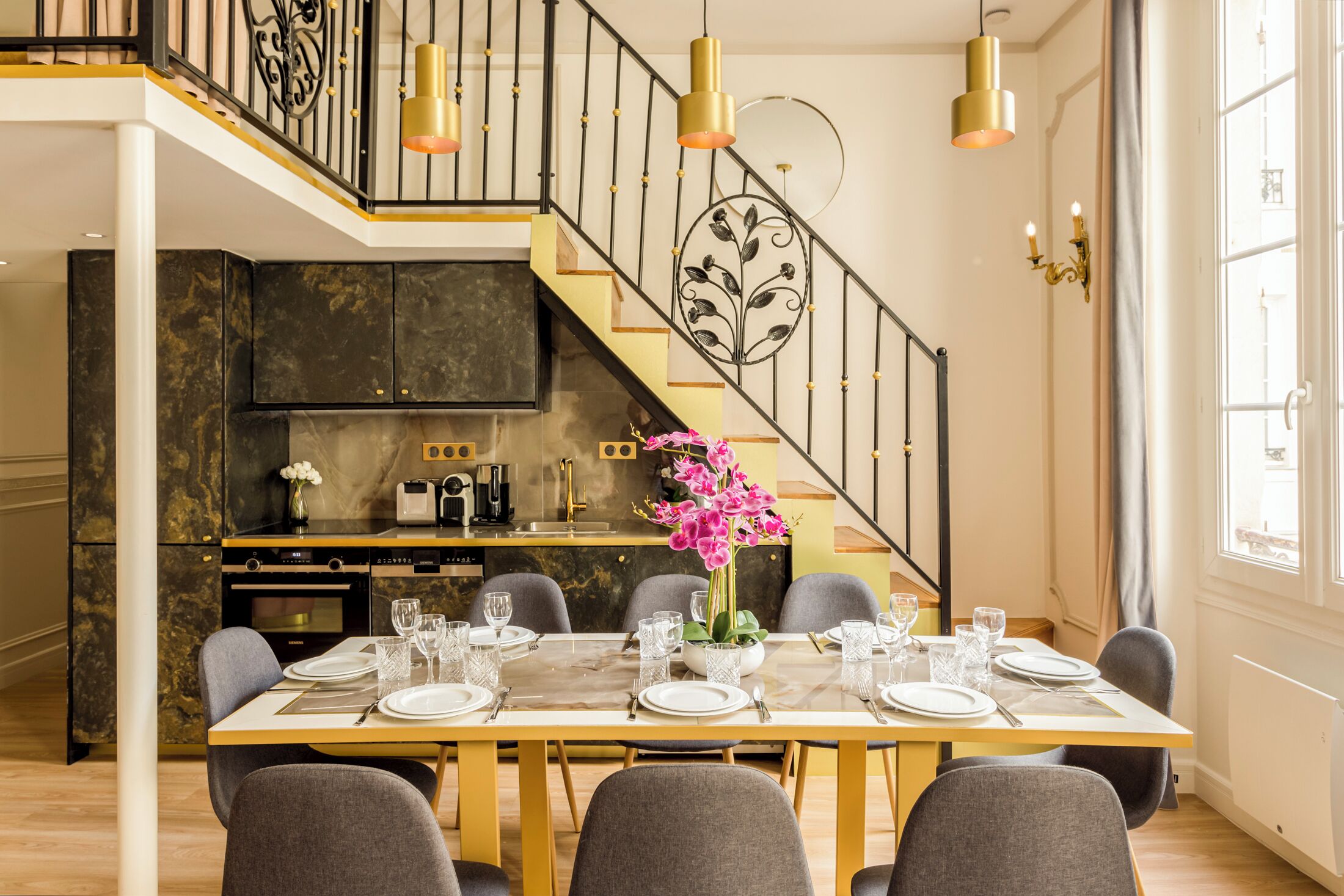 Un comedor con una mesa preparada para seis personas, una cocina al fondo y una escalera con barandilla.