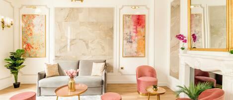 Un salon moderne avec un canapé gris, des fauteuils roses et une cheminée. Les murs sont ornés de tableaux abstraits et un lustre est suspendu au-dessus du canapé.