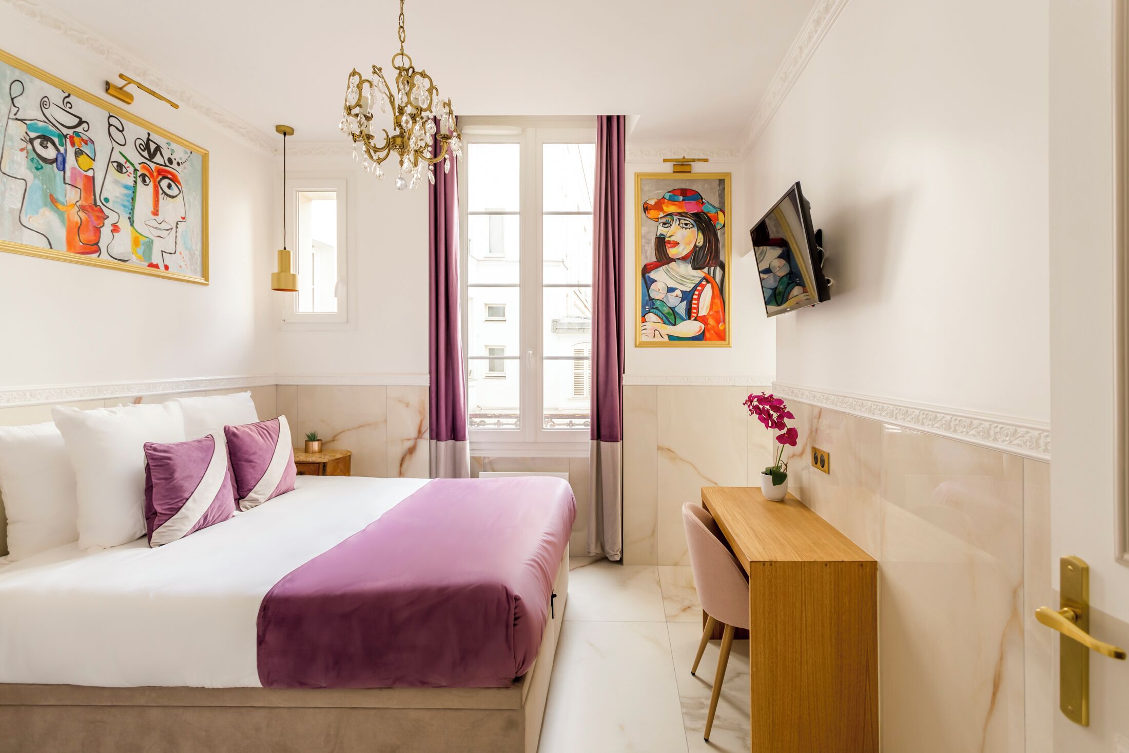 Habitación de hotel con cama, escritorio, silla, candelabro y obras de arte en las paredes.