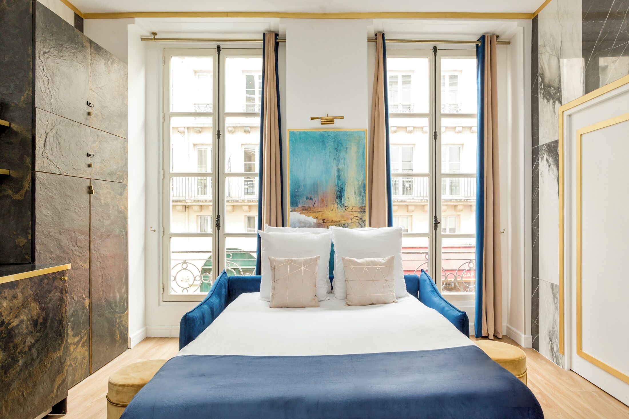 A bedroom with a bed, a large window, a painting, and a view of buildings.