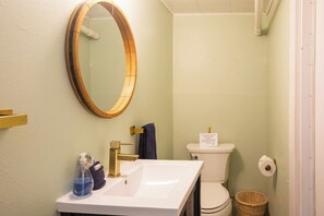A bathroom with a white sink, gold faucet, and a round mirror. There is a toilet with a paper towel holder and a basket. - Redwoods Oceanview Retreat (Elk)