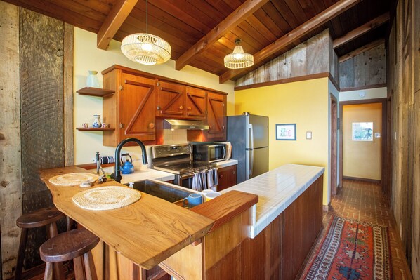 Una cocina con armarios de madera, un refrigerador de acero inoxidable, un microondas y una estufa. Hay dos taburetes de bar y un posavasos tejido en la encimera.