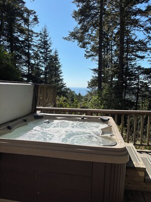 A hot tub on a wooden deck with a view of the ocean and tall trees.