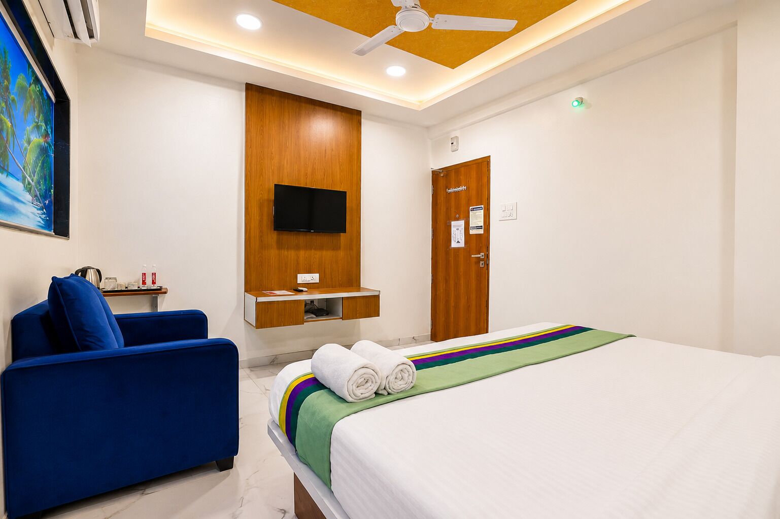 A hotel room with a bed, a blue sofa, a TV mounted on a wooden panel, and a ceiling fan.