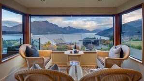 Un balcón con muebles de mimbre, una pequeña mesa con una botella y vasos, y una vista panorámica de montañas y un lago.