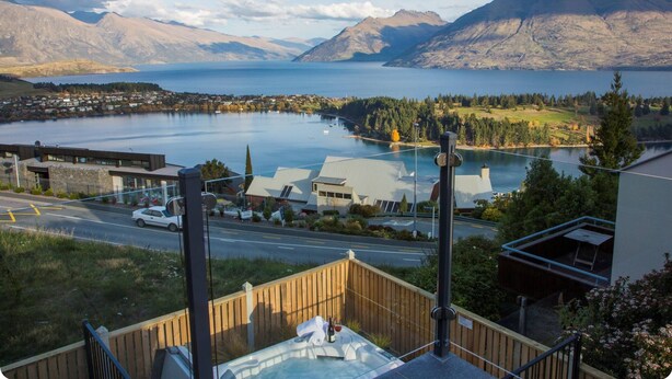 Vista panorámica de un lago con montañas al fondo, una carretera con un autom óvil y un edificio con terraza de madera en primer plano.