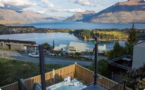 Una vista suggestiva di un lago con montagne sullo sfondo, una strada con un'auto, e un edificio con un terrazza in legno in primo piano.