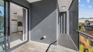 A modern balcony with a glass door leading to a kitchen, a metal railing, and a clear view of the surrounding neighborhood. - 2BR Apartment w/ Balcony Near Food Shop & Transit (Macgregor)