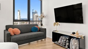A modern living room with a grey sofa, a flat-screen TV, and a view of city buildings. - Chic 1BR Getaway Steps from Beach&Surfers Paradise (Surfers Paradise)