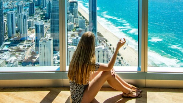 A woman sitting by a large window overlooking a beach and cityscape. - Chic 1BR Getaway Steps from Beach&Surfers Paradise (Surfers Paradise)