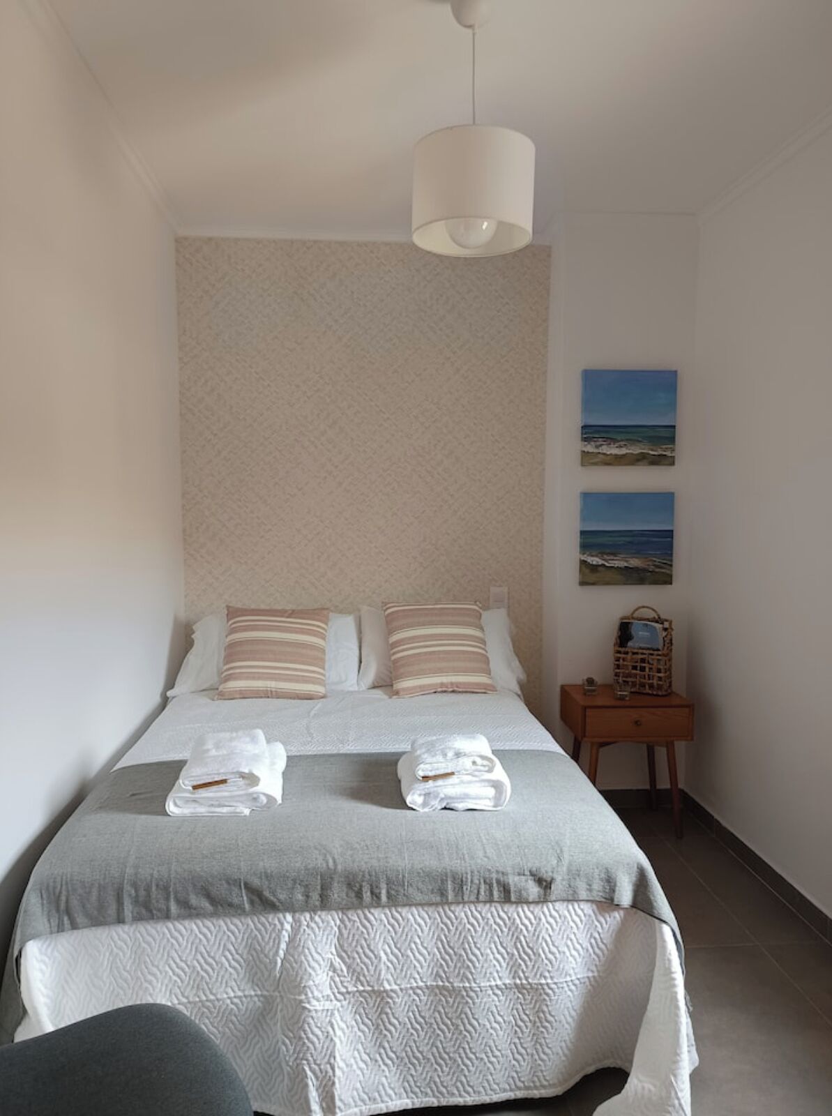 A neatly made bed with two pillows, a bedside table with a basket, and a wall-mounted painting of a beach scene.