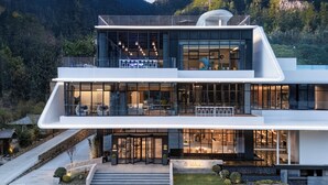 A modern hotel with a glass facade, outdoor seating, and a view of the surrounding mountains.
