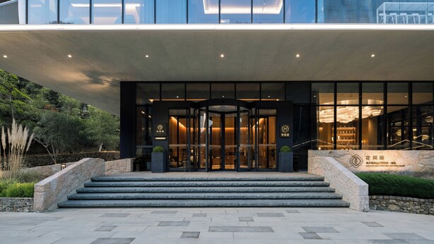 A modern hotel entrance with a glass facade, a set of stairs leading up to the doors, and a landscaped area with plants.
