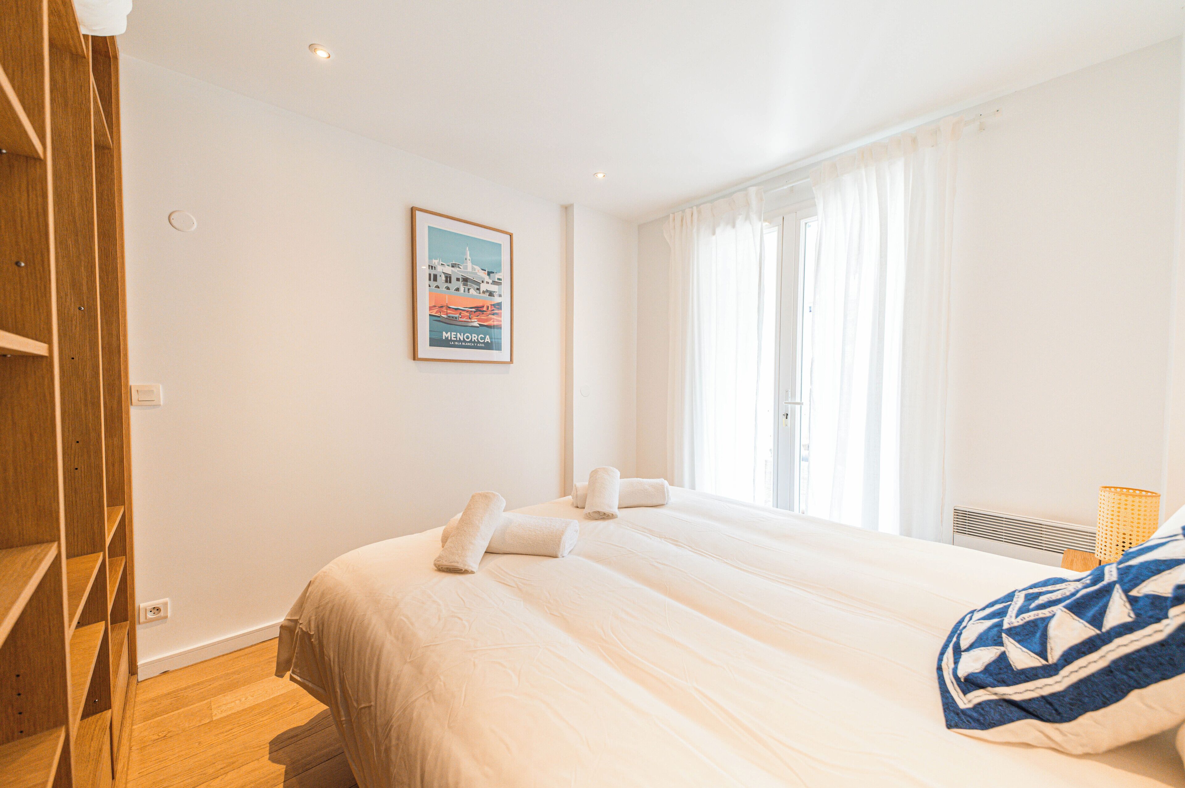 A hotel room with two beds, a wooden wardrobe, a framed picture on the wall, and a window with curtains.