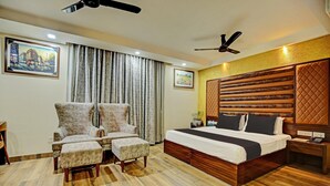 A bedroom with a large bed, two armchairs, a wooden headboard, ceiling fans, and a painting on the wall. - Deluxe Hotel Vihaan Residency (Dera Bassi)