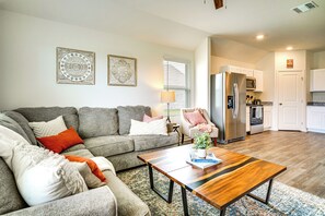 A living room with a grey sofa, a wooden coffee table, and a kitchen area with white cabinets and a stainless steel refrigerator.