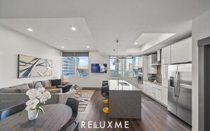 A modern apartment with a kitchen, dining area, and living room. It features a large window, a sofa, a dining table, and a kitchen island. - Reluxme | Uptown Lux High Rise Next to Stadiums🌇 (Charlotte)