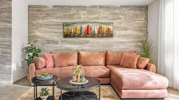 A living room with a sectional sofa, a round coffee table, a painting of sailboats, and a potted plant.