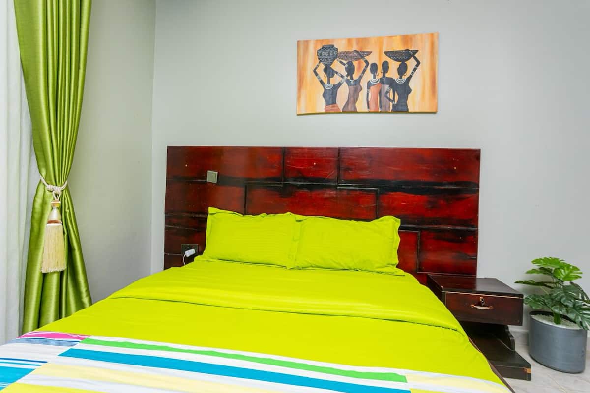 A bed with a bright green bedspread and pillows, a wooden headboard, a painting on the wall, a nightstand with a lamp, and a potted plant.