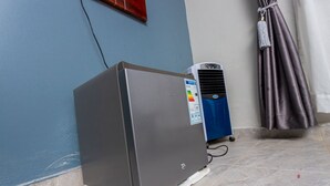 A compact refrigerator in a corner with a blue wall and a curtain. - The Den Soroti Green (Soroti)