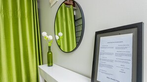 A bathroom with a green shower curtain, a small vase with white flowers, a round mirror, and a framed sign with house rules. - The Den Soroti Green (Soroti)