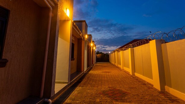 A corridor with a tiled floor leading to a building entrance, a barbed wire fence, and a clear sky. - The Den Soroti Green (Soroti)