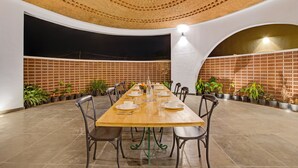 A long dining table set for a meal in a spacious, well-lit room with a high ceiling and brick walls. - ELIVAAS Urvi Resort | Eco-Luxury Getaway with Pool (Chikkellur)