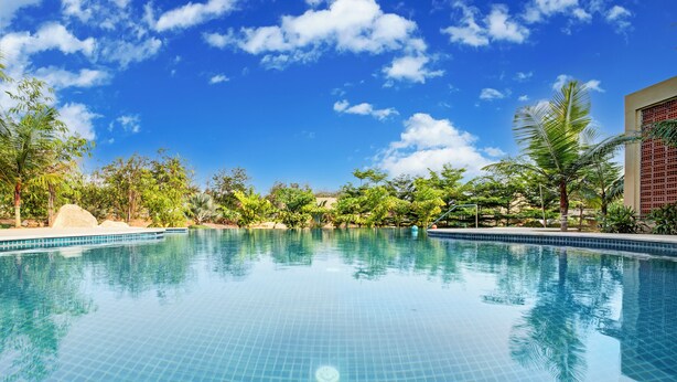 A clear blue swimming pool surrounded by tropical vegetation and a building with a tiled facade. - ELIVAAS Urvi Resort | Eco-Luxury Getaway with Pool (Chikkellur)