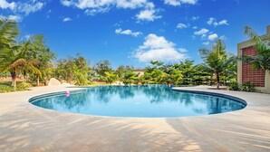 A large, curved swimming pool with a tiled edge, surrounded by palm trees and other greenery. - ELIVAAS Urvi Resort | Eco-Luxury Getaway with Pool (Chikkellur)