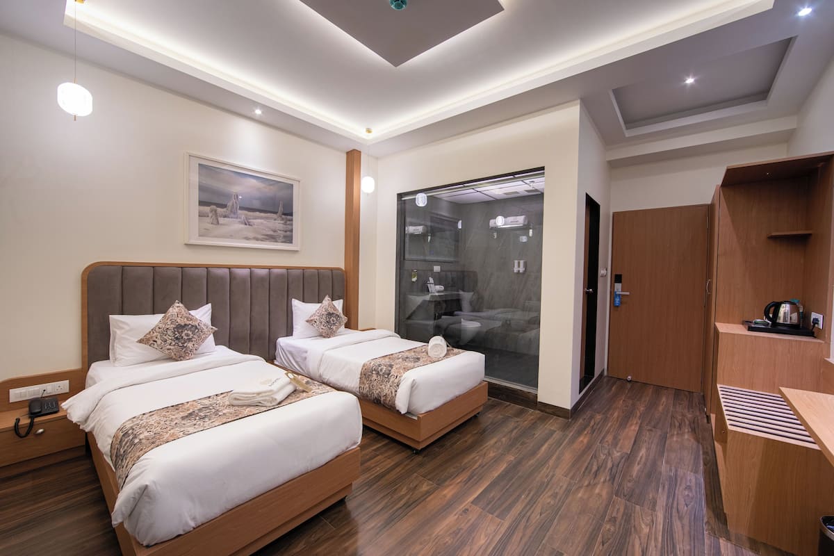 A hotel room with two beds, a large window, a wooden desk, and a bathroom visible through the glass door.