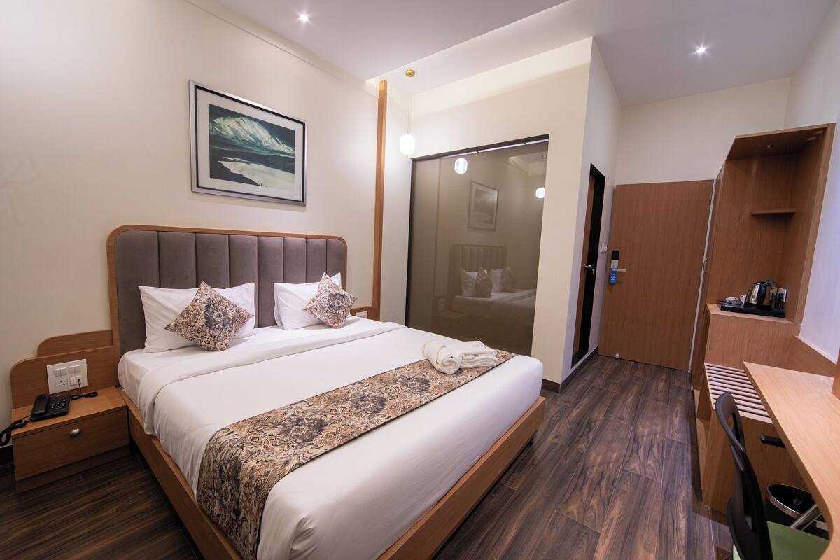 A hotel room with a large bed, a bedside telephone, a wooden nightstand, a mirror, a wardrobe, and a small desk with a chair.